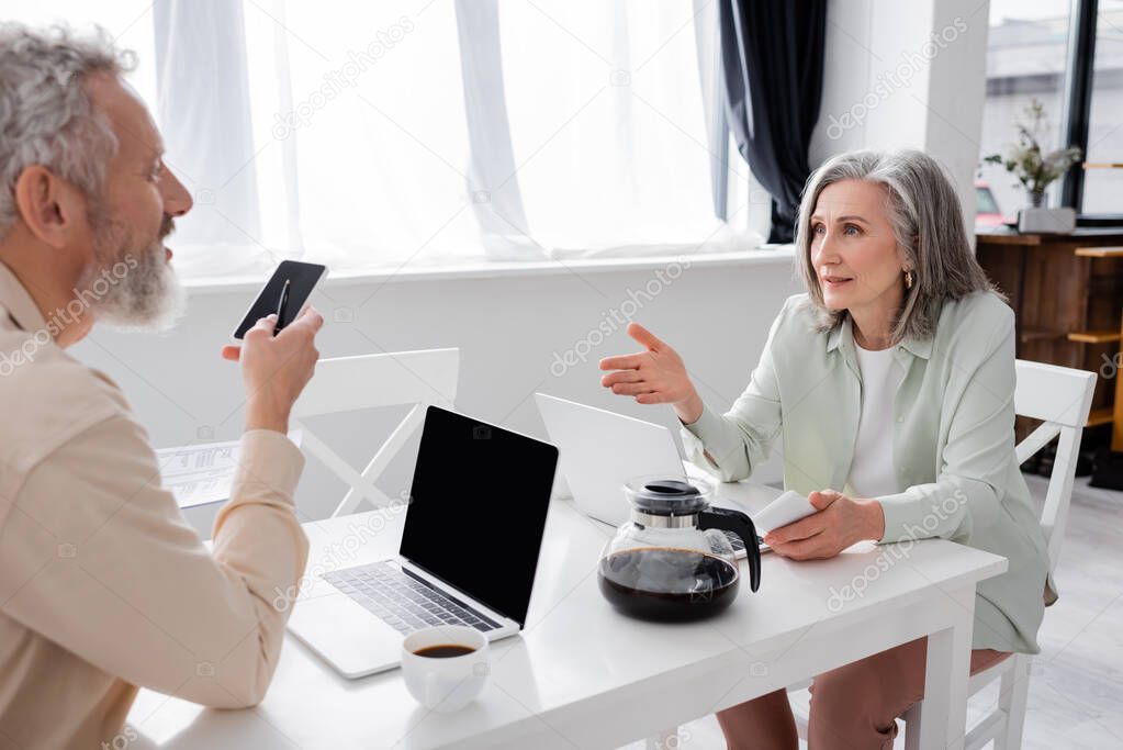 Mature woman with smartphone pointing with hand at husband with bills near laptops and coffee in kitchen 