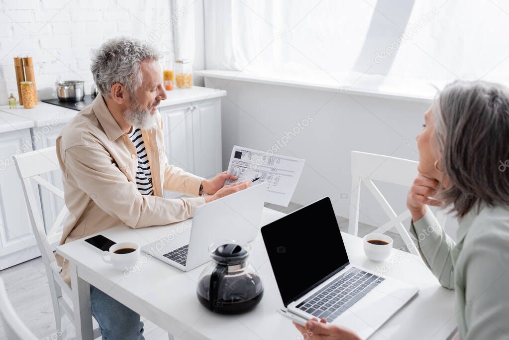 Mature man holding bills near devices and wife in kitchen 