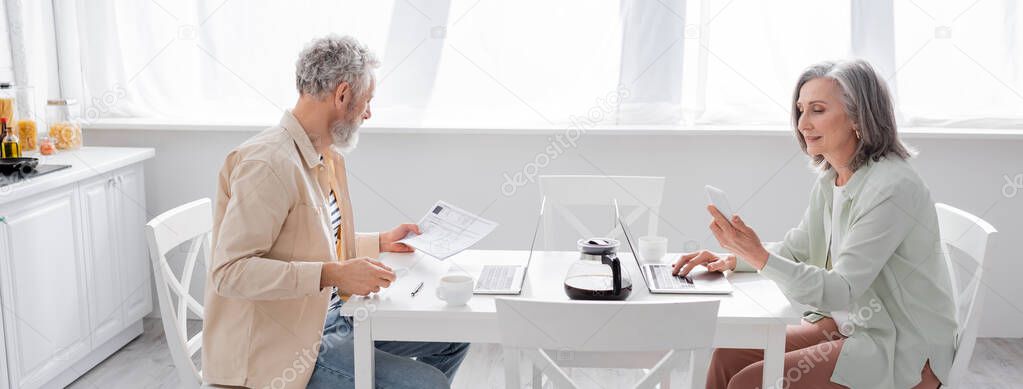 Mature couple counting bills near coffee and devices in kitchen, banner 