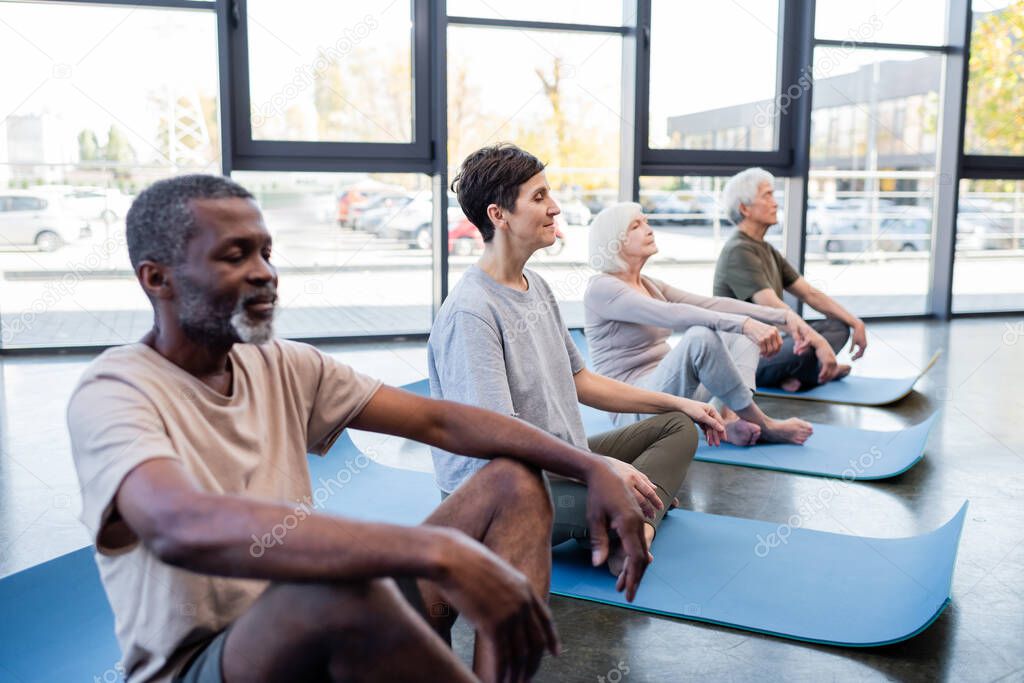 Senior woman meditating on yoga mat near interracial people in gym 