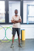 Senior afrikanisch-amerikanischer Sportler steht mit verschränkten Armen neben Fitnessmatte und Hula-Hoop-Reifen in Turnhalle 