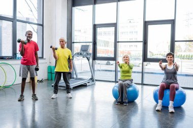 Multiethnic sportsmen training with dumbbells near sportswomen on fitness balls in gym  clipart
