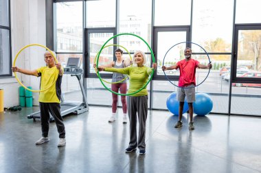 Çok ırklı son sınıf öğrencileri koşu bandının yanında hula çemberleri ve spor salonundaki fitness toplarıyla antrenman yapıyor. 
