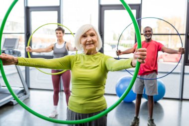 Senior woman smiling at camera while training with hula hoop in gym  clipart