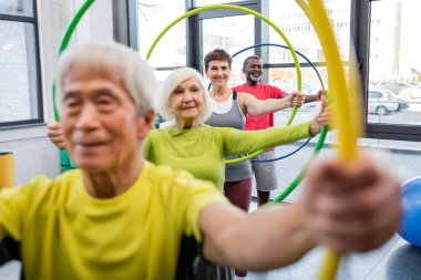 Smiling woman in sportswear training with hula hoop near multiethnic friends in sports center  clipart