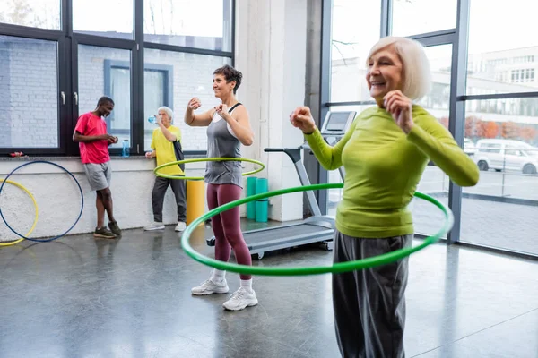 Yaşlı sporcu kadın, hula hoop ile spor salonunda arkadaş ve ırklar arası sporcular arasında antrenman yapıyor. 