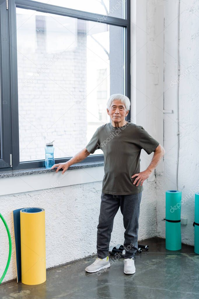 Elderly asian man looking at camera near fitness mats and sports bottle in gym 