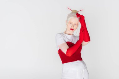 Young albino model in red gloves and hair sticks looking at camera isolated on white clipart