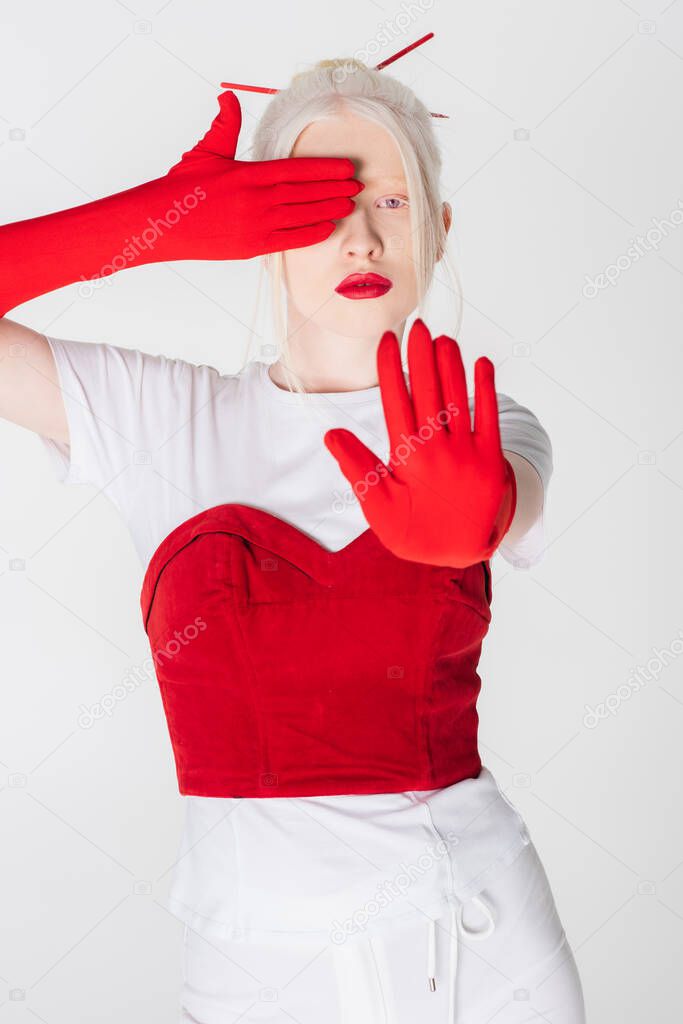Trendy albino model in gloves and red lips showing stop gesture isolated on white