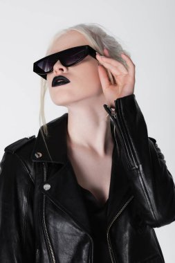Trendy albino woman in black leather jacket holding sunglasses isolated on white clipart