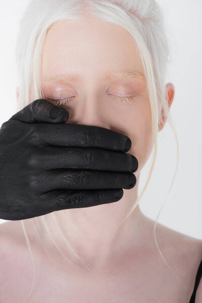 Male hand in black paint covering mouth of albino woman isolated on white