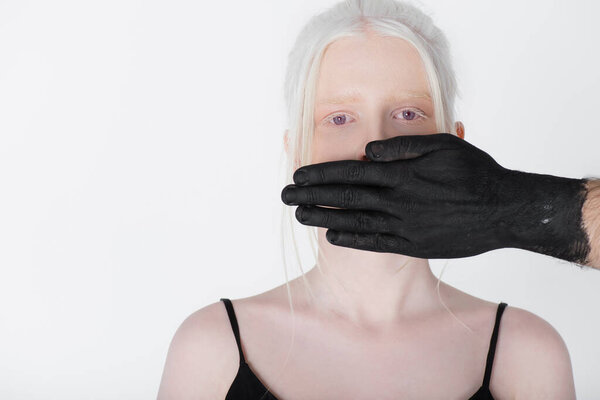 Male hand in black paint covering mouth of albino model isolated on white