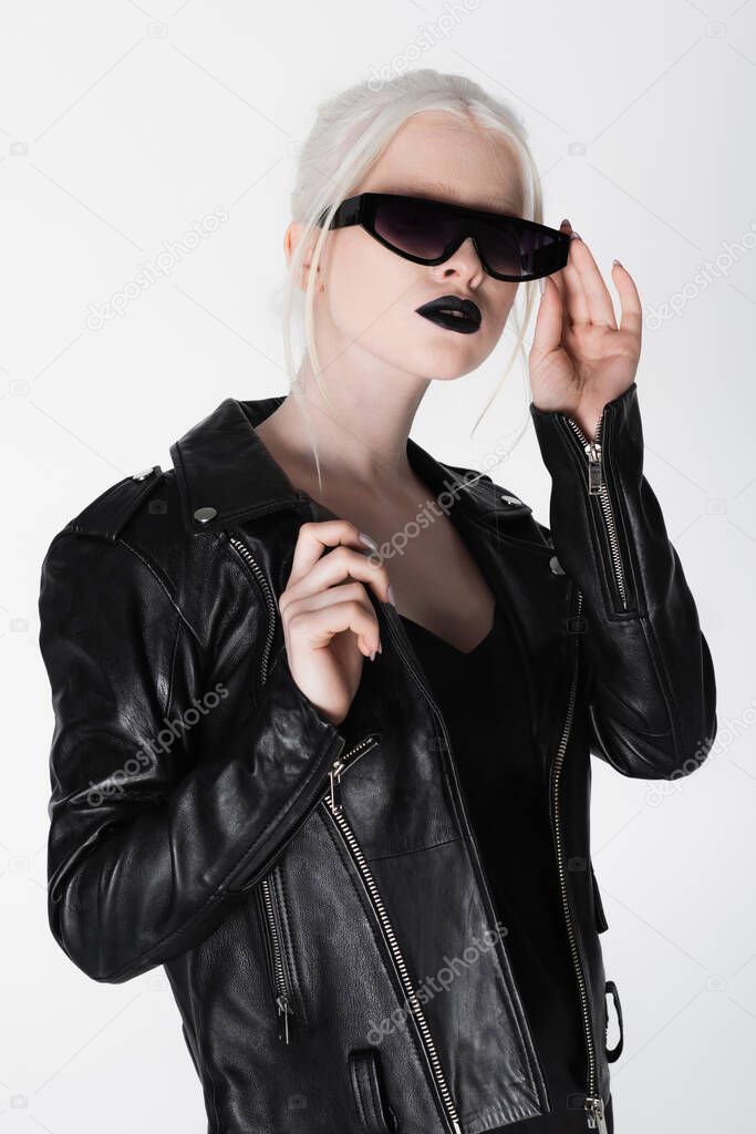 Stylish albino woman in leather jacket holding sunglasses isolated on white