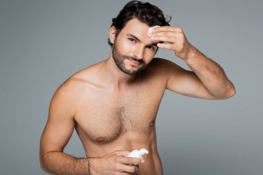 Mutlu gömleksiz adam yüzüne toner sürüyor ve pamuk pedi gri renkte izole edilmiş.