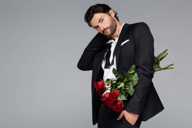 Kırmızı gül buketi tutan takım elbiseli sakallı adam elini cebine atmış gri renkte duruyor.