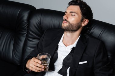 relaxed man in suit sitting on black sofa and holding glass of whiskey isolated on grey clipart
