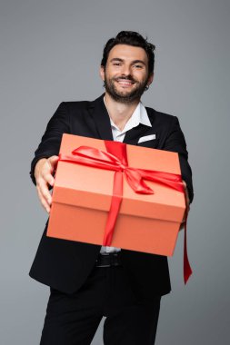 Takım elbiseli neşeli adam, gri üzerinde kırmızı hediye kutusu tutuyor. 