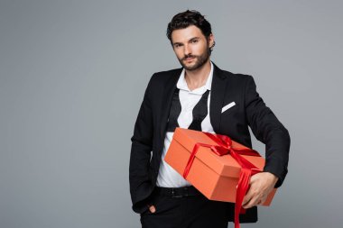 Takım elbiseli sakallı bir adam elinde kırmızı bir hediye kutusu ve elini cebine sokup gri renkte duruyor. 