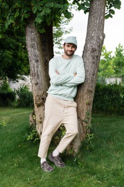 Tam boy sakallı adam çapraz kollu ve parktaki ağaç gövdelerine yaslanmış duruyor. 
