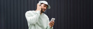 Beyzbol şapkalı sakallı adam metal çitlerin yanında kablosuz kulaklıkla müzik dinliyor.