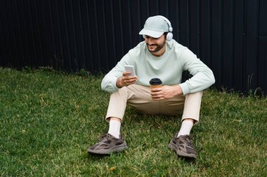 Kablosuz kulaklıkla müzik dinleyen ve çitin yanındaki yeşil çimlerde otururken cep telefonu kullanan olumlu adam.