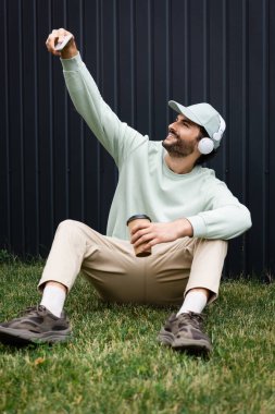 Şapkalı, kablosuz kulaklıklı, yeşil çimlerde kağıt bardakla otururken selfie çeken neşeli bir adam. 
