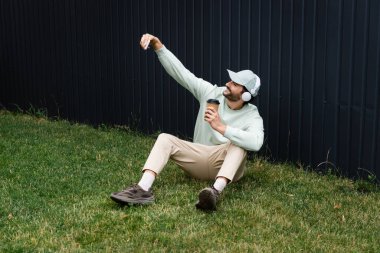 Kablosuz kulaklıklı mutlu adam yeşil çimlerde kağıt bardakla otururken selfie çekiyor. 