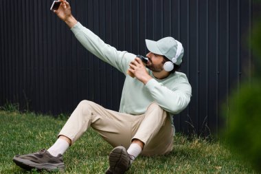 Kablosuz kulaklık takan, yeşil çimlerde elinde kağıt bardakla selfie çeken pozitif bir adam. 