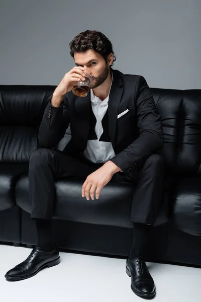 Młody Brodaty Mężczyzna Formalnym Ubraniu Siedzi Czarnej Sofie Pije Whisky — Zdjęcie stockowe