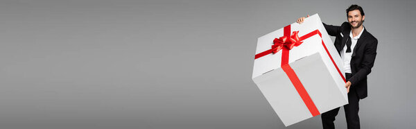 счастливый человек в костюме держит огромный подарочный коробок изолирован на серый, баннер