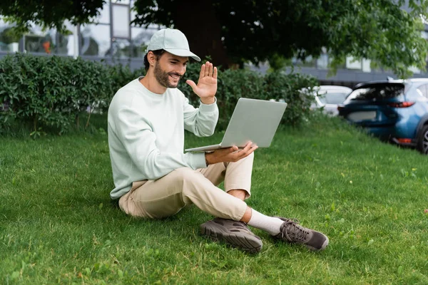 full length of joyful man waving hand during video call while sitting with crossed legs on lawn