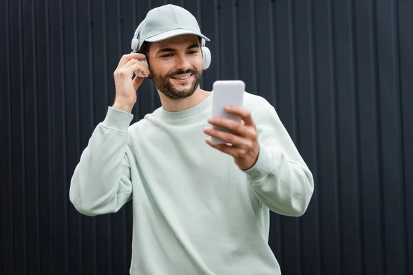 戴便帽的男人在无线耳机里听音乐 在金属栅栏附近使用手机 — 图库照片
