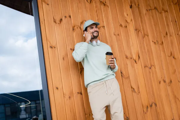 Szczęśliwy Człowiek Czapce Trzymając Kawę Iść Podczas Rozmowy Smartfonie Pobliżu — Zdjęcie stockowe