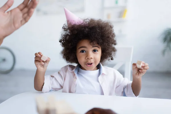 excited african american girl in party cap looking at camera while holding wooden blocks