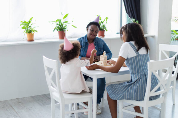 african american child with mom and granny in party caps playing wood blocks game at home