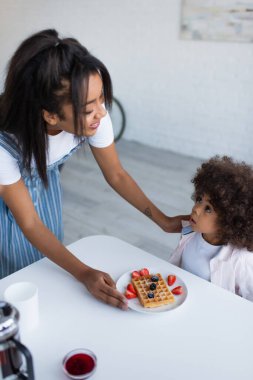 Afro-Amerikan kızı gülümseyen annesine bakıyor kahvaltıda taze böğürtlenli waffle servis ediyor. 