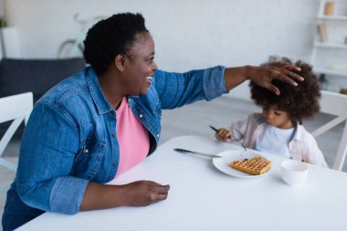 Mutlu Afro-Amerikan büyükanne kahvaltıda waffle yiyen torunun kıvırcık saçlarına dokunuyor.