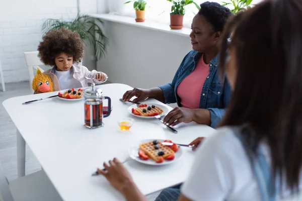 アフリカ系アメリカ人の女の子は柔らかいおもちゃでテーブルに座っている間におばあちゃんとぼやけたお母さんと朝食を持っています — ストック写真
