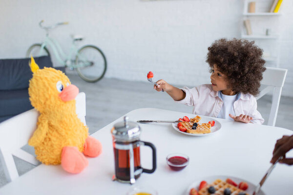 african american girl proposing strawberry to soft toy while having breakfast in kitchen