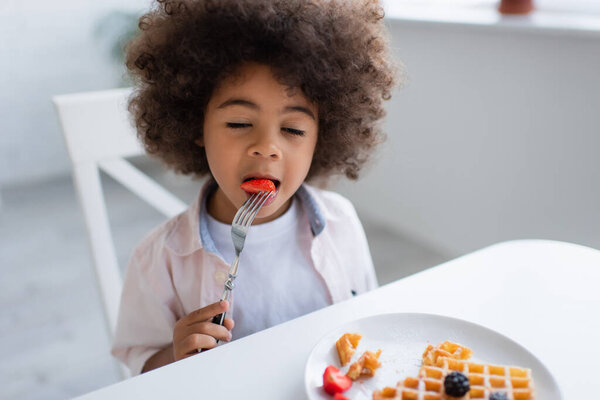 curly african american girl with closed eyes eating strawberry near tasty waffle on plate