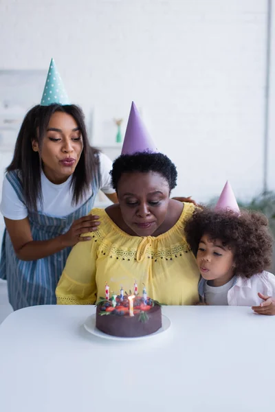 非洲裔美国妇女和戴着派对帽的蹒跚学步的女孩吹灭生日蛋糕上的蜡烛 — 图库照片