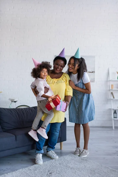 非洲裔美国妇女和儿童头戴党帽 一边拿着礼物 一边在客厅对着相机微笑 — 图库照片