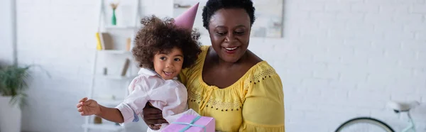 快乐的中年非洲裔美国女人 头戴党帽 头戴礼品盒 打着横幅 抱着孙女 — 图库照片