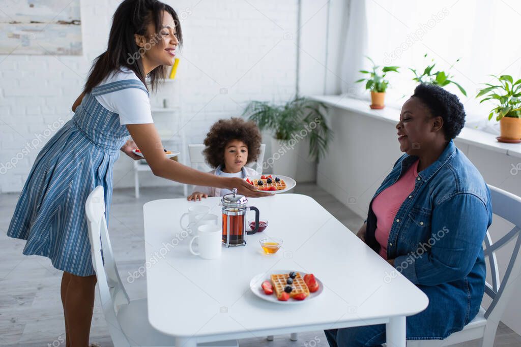 smiling african american woman holding plate with waffle and berries near mom and daughter