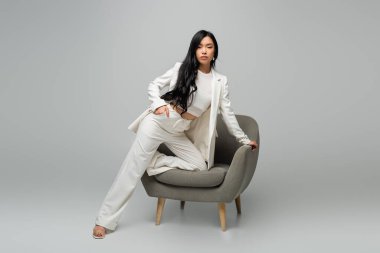 full length of young asian woman leaning on armchair while posing with hand in pocket on grey clipart