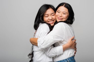Beyaz gömlekli mutlu Asyalı anne ve kız gri gömlekli izole edilmiş bir şekilde sarılıyorlar.