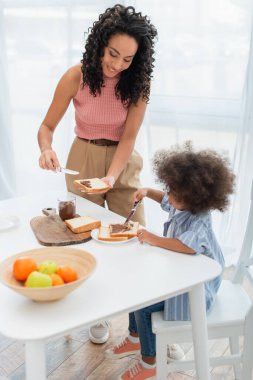 Afro-Amerikan anne meyvelerin yanında ekmeğin üzerine çikolata ezmesi döküyor ve çocuk mutfakta. 