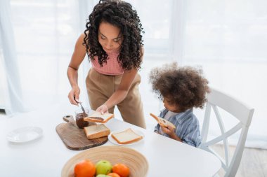 Genç Afrikalı Amerikalı anne çocuğun yanına çikolata ezmesi sürüyor ve mutfakta meyveler var. 
