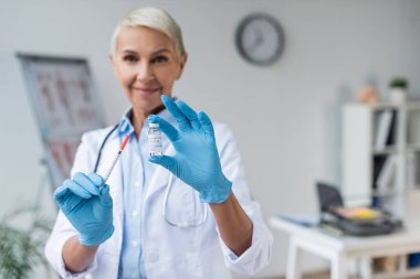 Ellerinde Coronavirus aşısı ve şırıngayla şişe tutan lateks eldivenli bulanık ve gülümseyen bir doktor.