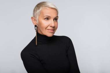 Uzun küpe ve balıkçı yaka kazak giyen neşeli, son sınıf öğrencisi bir kadın griye tek başına bakıyor.